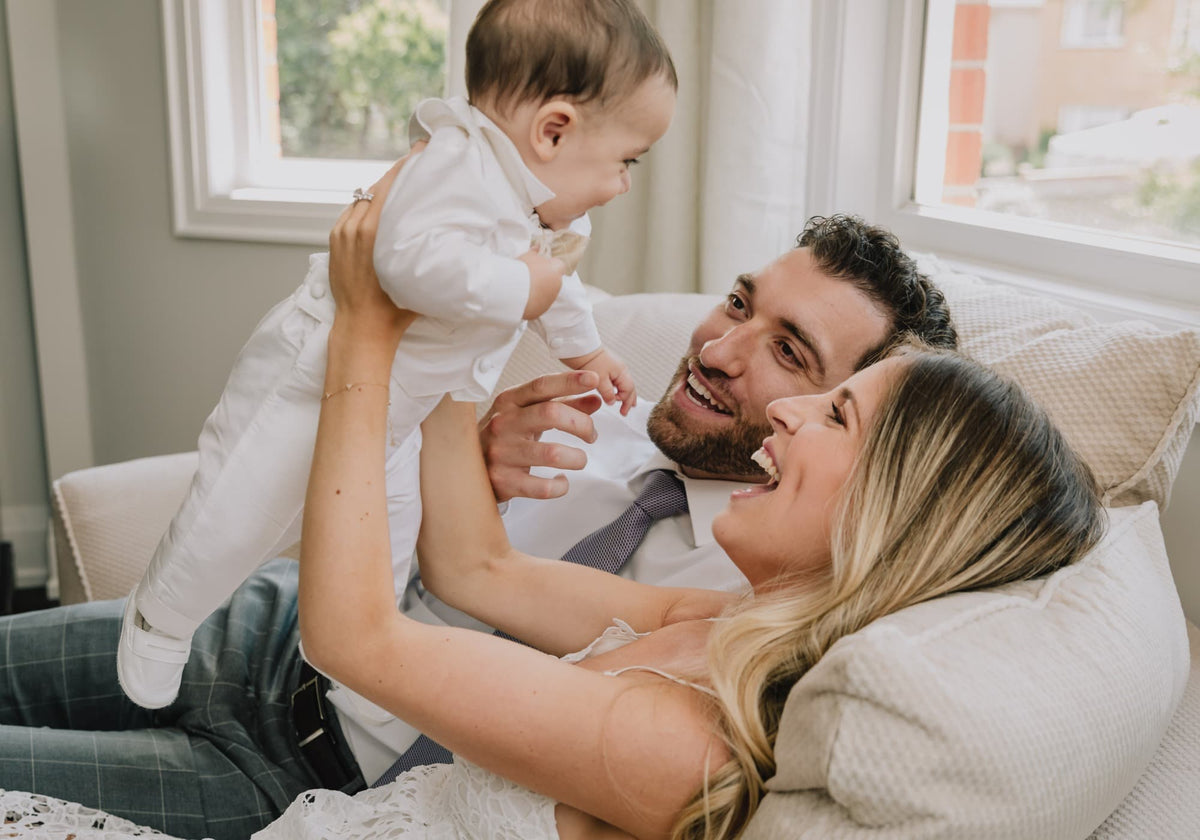 Familie mit Baby auf dem Arm, Eltern lachen im Wohnzimmer, Kind spielt und wirkt fröhlich
